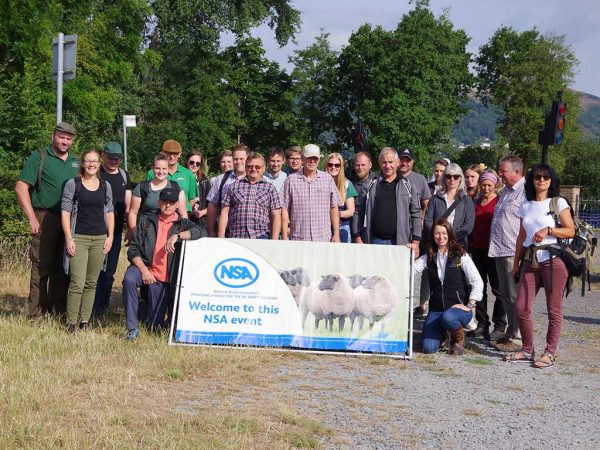 Eine Gruppe von Menschen steht zusammen. Im Vordergrund ist ein Banner, auf dem Schafe zu sehen sind und der Text „Welcome to this NSA event“ geschrieben ist. Im Hintergrund ist eine Berglandschaft und grüne Bäume zu sehen.