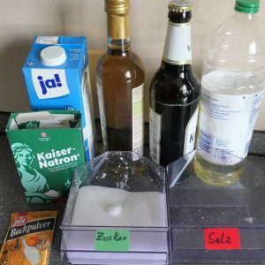 Tüte mit Backpulver, 2 Behälter mit jeweils Zucker und Salz, im Hintergrund: eine Packung Kaiser-Natron und 3 verschiedene Flaschen (Essig, Bier, Öl)