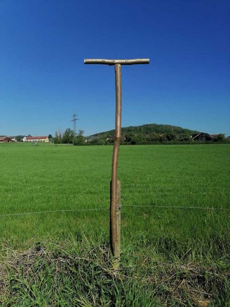 Rundholz mit Querbalken auf zwei Meter Höhe. Im Hintergrund eine grüne Wiese, landwirtschaftliche Gebäude und ein bewaldeter Hügel