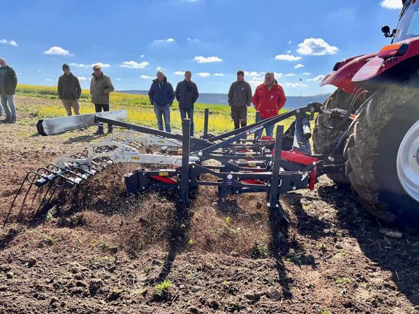 Zu sehen in das drei Meter breite Anbaugerät auf einem unbestellten Feld, im Hintergrund einige Personen.