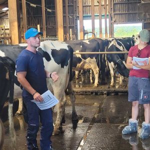 Landwirt in einem Kuhstall erklärt einer Gruppe von Azubis, was bei einer Tierbeurteilung zu beachten ist; links und im Hintergrund sind die Kühe zu sehen; die Personen halten Collegeblocks in der Hand