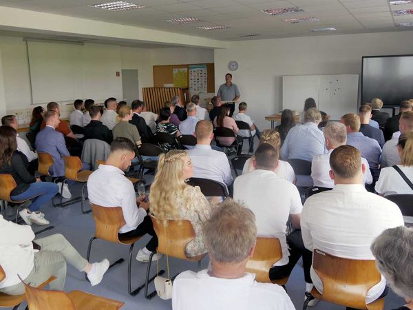 Personengruppe sitzt in einem Raum auf Stuhlreihen und hört einem Redner zu