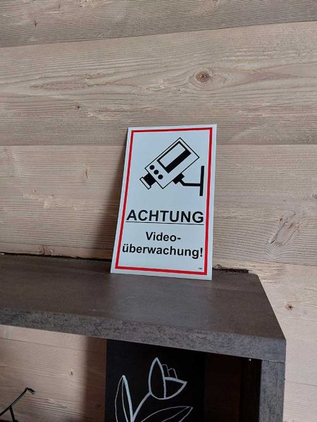 Das Bild zeigt ein rot-weißes Schild auf einem Regal, welches eine Kamera abgebildet hat und die Worte „Achtung Videoüberwachung“ trägt