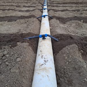 Ein weißer Feuerwehrschlauch liegt in gerader Linie auf dem Acker. In regelmäßigen Abständen (75cm) ragen T-Stücke aus dem Schlauch. Auf beiden Seiten sind Tropfschläuche angeschlossen die nach wenigen Zentimetern in der Erde verschwinden. Dunkle Streifen auf der Ackeroberfläche sind entlang der vergrabenen Tropfschläuche deutlich zu erkennen. Im Hintergrund verläuft eine Straße auf dieser sind zwei Fahrradfahrer zu sehen.