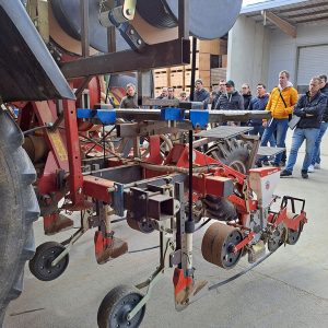 Das Stück eines Traktorhinterrads ist an der linken Seite zu sehen. An dem Traktor hängt eine rote Sämaschine. Über der Sämaschine hängen zwei erkennbare Rollen mit Tropfschläuchen. Von den Rollen wird der Tropfschlauch durch ein Rohr hinter ein Verlege-Schar geführt. Die Maschine wird von etwa 20 Menschen im Hintergrund begutachtet.