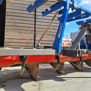 Ein roter Dammformer steht in einem Hof im Hintergrund stapeln sich Holzkisten. Der Dammformer ist mit schwarzen Verlegescharen und blauer Abrollvorrichtung ausgestattet.