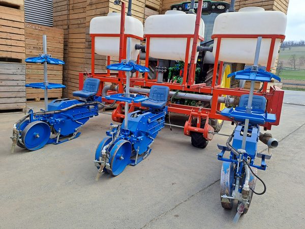 Pflanzmaschine und Traktor stehen in einem Hof mit Holzkisten im Hintergrund. Es sind drei blaue Pflanzstationen mit Sitz zu sehen. Dahinter befinden sich 3 weiße Wassertanks in einem roten Metallgestänge.