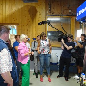 Eine Gruppe Personen begutachtet einen Melkroboter im Stall. Rechts hinten ist ein Filmteam mit Kamera und Mikrofon zu sehen.