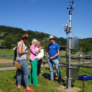 Mehrere Personen besichtigen eine Wetterstation. Im Hintergrund ist Wald zu sehen, der Himmel ist blau.