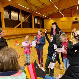 In einer Reithalle stehen mehrere Kinder und Erwachsene in einem Kreis. Die Kinder halten Zettel in den Händen und scheinen auf Fragen zu antworten,