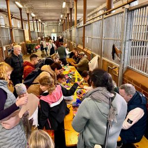 In einer großen Halle mit Pferdeboxen, dem Stall III des Landgestüts, sitzen an den Seiten viele Kinder und Erwachsene an langen Holztischen. Sie basteln mit buntem Papier und Materialien ihre neuen Steckenpferde, während einige Erwachsene den Kindern helfen.