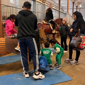 Auf dem Bild sind mehrere Personen im sogenannten Stall III des Landgestüts zu sehen, eine große, längliche Halle mit Einzelboxen für Pferde. Im Vordergrund sitzt ein Kind in einer pinken Jacke auf einem Holzpferd, während ein Erwachsener, offensichtlich der Vater, sich daneben befindet und mit dem Kind interagiert. Zwei Kinder in grünen Trainingsanzügen spielen auf einem blauen Schaukelpferd in der Nähe.