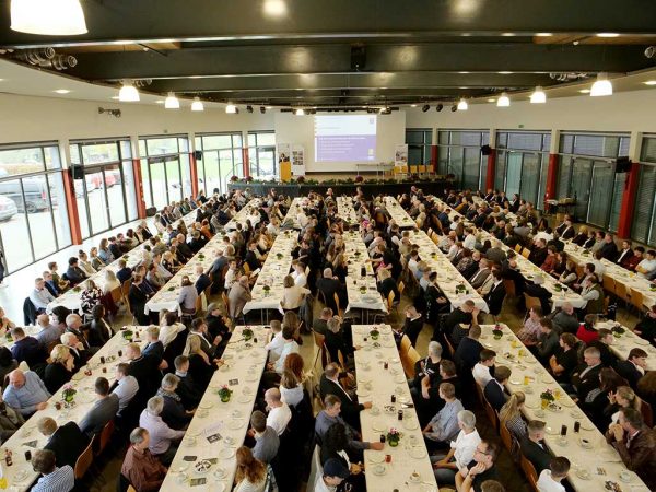 Blick in eine Halle mit Tischen und Stühlen, an denen Menschen sitzen; im Hintergrund eine Bühne mit Rednerpult