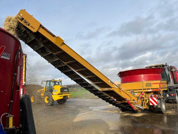 Über ein Förderband fließt zerkleinertes Stroh aus einer Strohmühle (rechts) in einen Futtermischwagen. Im Hintergrund steht ein Radlader.