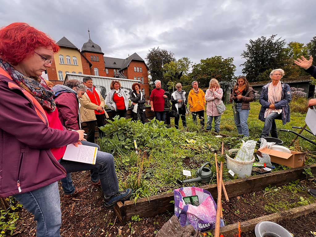 Personengruppe steht an einem Gemüsebeet ; im Vordergrund sieht man Gartengeräte, eine Gießkanne und einen Sack mit Blumenerde