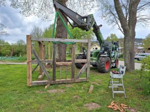 Eine Holzständerkonstruktion wird auf einer Wiese mit einem Gurt von einem Teleskoplader angehoben. Im Vordergrund steht eine kleine Leiter mit Werkzeugkoffer, daneben liegen Spanngurte. Im Hintergrund sind Bäume, ein Gehweg sowie Gebäude und parkende Autos zu sehen.