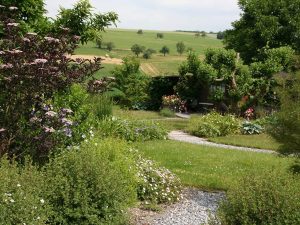 Garten mit Rasen- und Wiesenflächen, Büschen Stauden und Wegen; im Hintergrund hügelige Landschaft