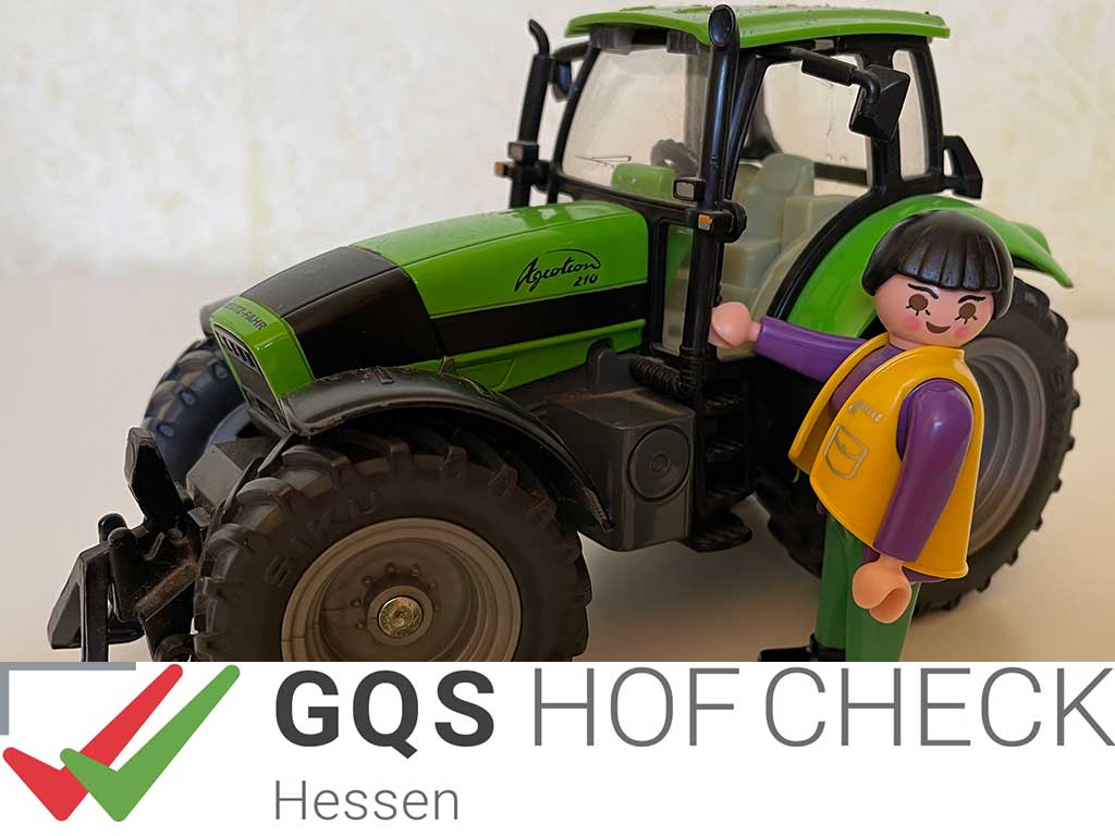 Spielzeugtrecker und Playmobilfigur; im Vordergrund Logo: GQS HOFCHECK Hessen