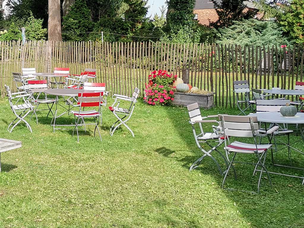 ländliches Gartenlokal auf einem Rasen; zu sehen sind 3 leere Tische mit Klappstühlen; im Hintergrund Blumen und ein Staketenzaun