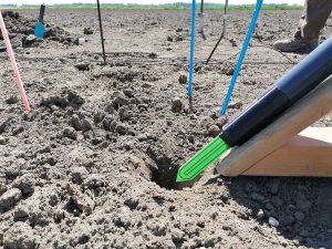 Im Vordergrund ist ein Holzgestell zu sehen, das als Winkelhilfe für einen Erdbohrer dient. Es ermöglicht das Bohren von Löchern im 45-Grad-Winkel. Auf dem Gestell liegt ein Einbaurohr, an dessen vorderem Ende ein grüner SMT-100-Bodensensor mit einer markanten, dreieckigen Sensorspitze befestigt ist. Auf der sichtbaren Messfläche des Sensors befinden sich zwei U-förmige Drähte, die zur Messung der Bodenfeuchte dienen. Direkt vor dem Sensor befindet sich ein kleines, schräg in den Boden gebohrtes Loch – es ist exakt auf den Durchmesser des Sensors abgestimmt und dient als Einführöffnung. Im Hintergrund des Bildes sind verschiedenfarbige Kunststoffstäbe, Sensorkabel und diverse Werkzeuge zu erkennen, die auf die laufende Installation im Versuchsfeld hinweisen.