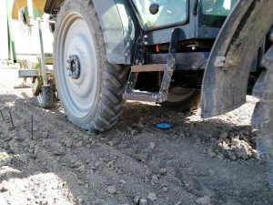 Ein Traktor fährt über einen unbewachsenen Acker. Unter dem Fahrzeug befindet sich ein kleiner, blauer Kanaldeckel mit einer schwarzen, quadratischen Einfassung, der bündig im Boden eingelassen ist. Am linken Bildrand sind drei schwarze Kunststoffstäbe im Boden zu sehen –zur Markierung von Messpunkten.