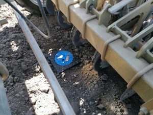 Eine Drillmaschine befindet sich in Fahrtrichtung direkt vor einem kleinen, blauen Kunststoffkanaldeckel mit schwarzer, quadratischer Einfassung im Boden. Die Maschine ist kurz davor, diesen zu überqueren. Der Kanaldeckel ist bündig im unbewachsenen Ackerboden eingelassen.