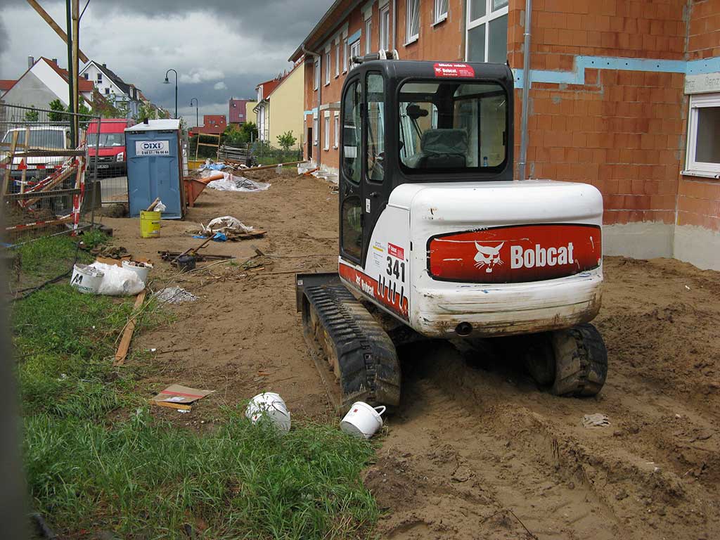 Schwere Baumaschinen verdichten den Boden. Neben einem unverputzten Neubau steht ein Bagger auf dem feuchten Erdboden. Im Boden sind tiefe Fahrspuren erkennbar.