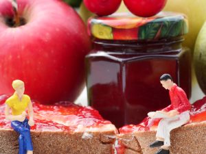 Zwei Miniaturfiguren sitzen auf einem mit Fruchtaufstrich beschmierten Brot. Dahinter ist ein roter reifer Apfel sowie ein Glas mit rotem Fruchtaufstrich zu sehen.