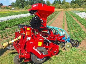 Eine rote Maschine zur flachen Bodenbearbeitung steht vor Gemüsebeeten, im Hintergrund sind Bäume zu sehen.
