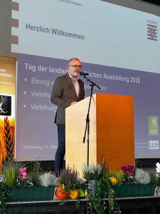 Redner steht an einem Pult mit Mikrofon; im Vordergrund herstliche Blumendekoration; im Hintergrund eine Präsentationsleinwand mit Text "Herzlich willkommen"