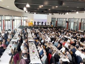 Blick in einen Veranstaltungssaal mit Tischreihen, an denen Personen sitzen; vorne eine Bühne mit 5 Musikern