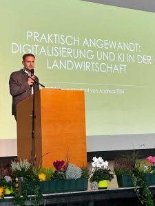 Redner steht an einem Pult mit Mikrofon; im Vordergrund herstliche Blumendekoration; im Hintergrund eine Präsentationsleinwand