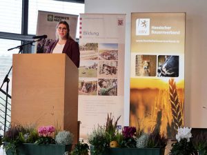 Rednerin steht an einem Pult mit Mikrofon; im Vordergrund herstliche Blumendekoration; im Hintergrund verschiedene RollUp's Präsentationsleinwand mit Text "Herzlich willkommen"