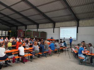 Das Foto zeigt Besucherinnen und Besucher in einer Halle. Sie sitzen auf Bierbänken und hören einem Vortrag zu. Der Referent steht mit Mikrofon vor einer Leinwand.