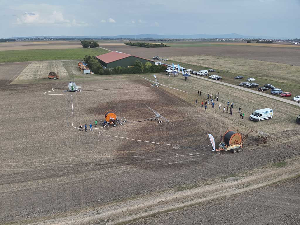 Das Foto zeigt aus der Luft die Vorführung von Bewässerungsmaschinen. Im Hintergrund ist die Maschinenhalle, daneben Besucher und parkende Autos auf einer Wiese.