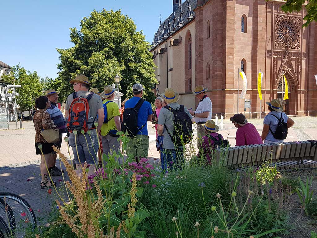 Personengruppe mit Strohhüten auf den köpfen steht voe einem Backsteingebäude; im Vordergrund ein blühendes Blumenbeet und eine Holzbank