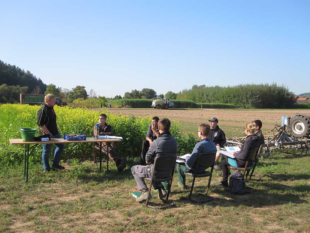 Eine Gruppe Menschen sitzt auf Stühlen, ein Ausbilder steht hinter einem Tisch, dahinter ist ein blühendes Rapsfeld.