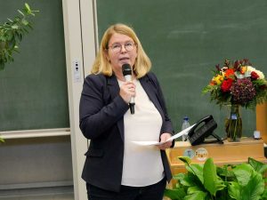 Rednerin mit Mikrofon; im Hintergrund Blumen