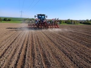 Ein Traktor zieht ein mehrreihiges Hackgerät über ein frisch bepflanztes Feld, im Hintergrund sind Grünflächen zu sehen, während das Gerät beim Arbeiten Staub aufwirbelt.