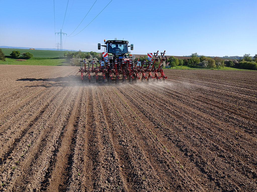 Ein Traktor zieht ein mehrreihiges Hackgerät über ein frisch bepflanztes Feld, im Hintergrund sind Grünflächen zu sehen, während das Gerät beim Arbeiten Staub aufwirbelt.