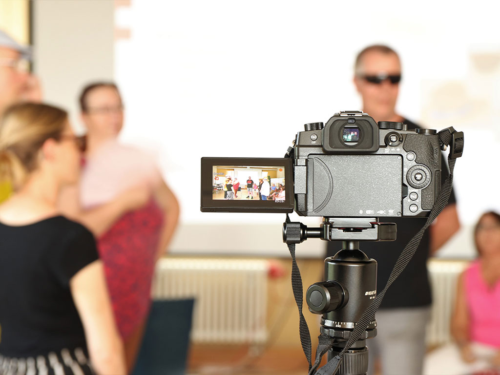 Im Vordergrund steht eine Kamera, die auf vier Laienschauspieler gerichtet ist, die ein Rollenspiel durchführen.