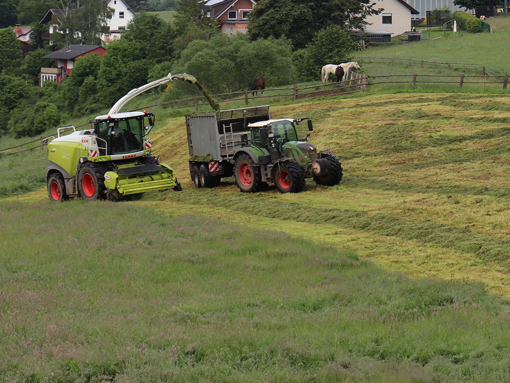 Zwei landwirtschaftliche Maschinen auf einer Wiese, im Hintergrund Bäume und Wohnbebauung