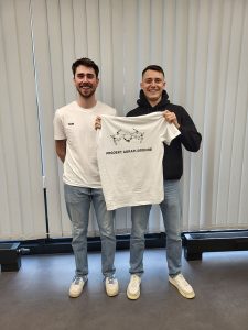 2 männliche Personen stehen in einem Raum und halten gemeinsam ein weißes T-Shirt mir dem Aufdruck "Projekt Agrar-Drohne" in den Händen