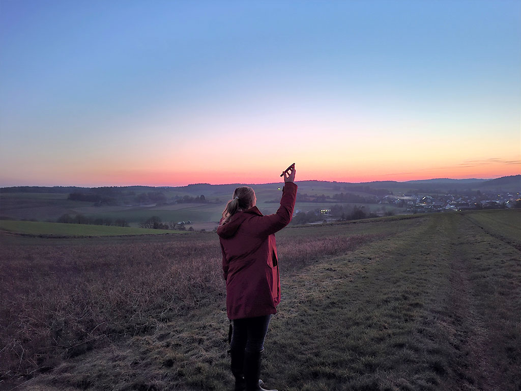Eine Frau steht bei Sonnenaufgang auf einer Rebhuhn-Fläche und hält eine Klangattrappe in der Hand.