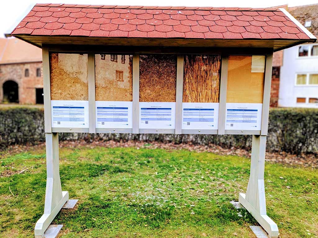 Rückseite einer beidseitig begehbaren Ausstellungswand mit Dämmstoff-Exponaten. Präsentiert werden Holzfaser, Schilf, Stroh, Hanf und Hanfjute, jeweils mit kurzen Informationen zu Eigenschaften und Einsatzmöglichkeiten.