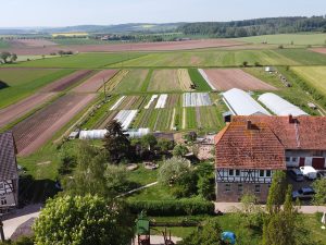 Drohnenaufnahme des Betriebsgeländes. Zu sehen ist die Hofstelle, ein Garten und dahinterliegende Gemüsefelder mit Folientunneln. Der Blick in die Ferne ist von Feldern und Bäumen geprägt.