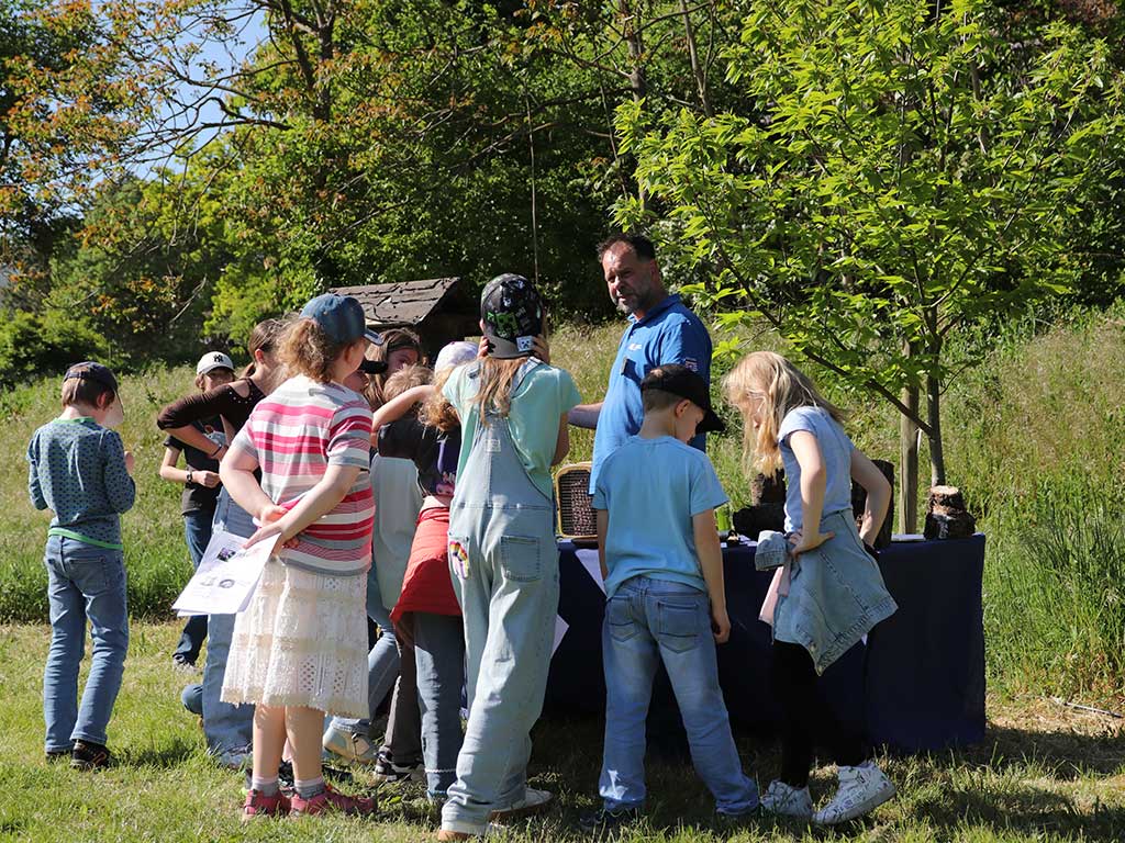Eine Kindergruppe steht im Freien um einen Tisch mit Materialien, die Wildbienen als Nisthilfe dienen können und hören dem Referenten zu, der den Einsatz der Materialien erklärt.