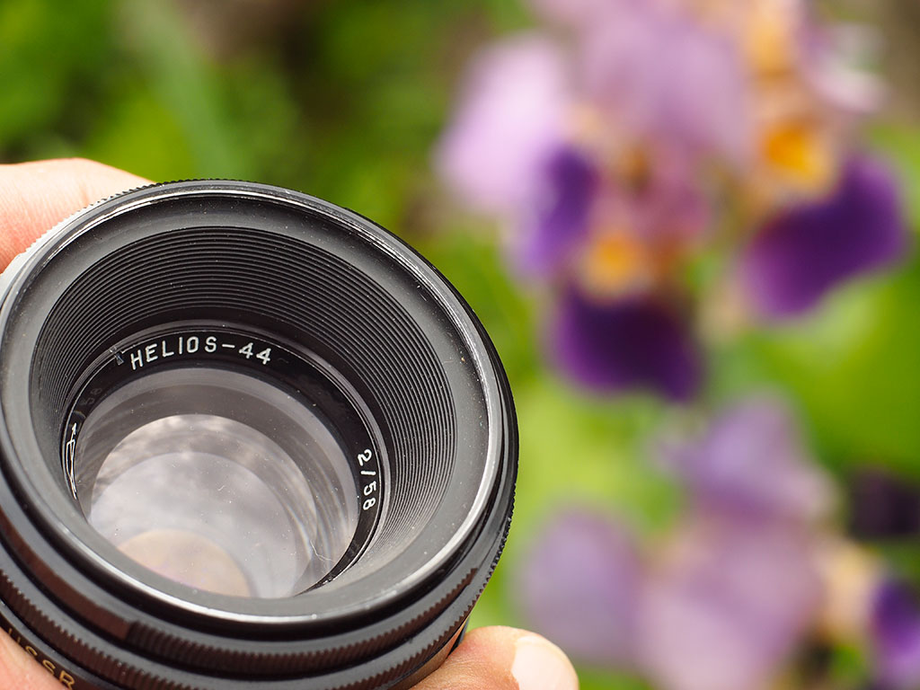 Das Foto zeigt ein altes analoges Objektiv, welches in einer Hand gehalten wird. Im Hintergrundbokeh sind Gartenblumen zu erahnen.