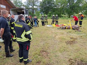 ein Mann demonstriert an einem Pferdedummy das Anlegen von Rettungsgurten; ein Feuerwehrmann steht daneben; eine Personengruppe schaut zu