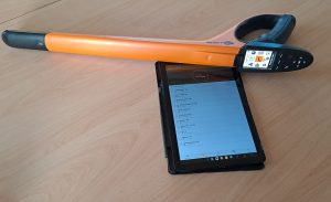 Ein revolverförmiges Ohrmarkenlesegerät und rechteckigem buntem Display oberhalb des Griffes liegt auf einem Tisch neben einem Tablet auf dessen Bildschirm ein Dateiverzeichnis zu erkennen ist.